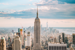 A far away view of New York City with the Empire State building prominent in the centre against a cloudy sky with the sun behind the clouds