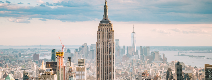 A far away view of New York City with the Empire State building prominent in the centre against a cloudy sky with the sun behind the clouds