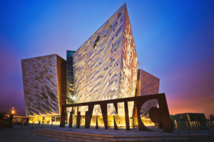 A ground level photo of the Belfast Titanic building during ealry evening, lit from the base with a dark blue sky and the sun setting behind with a little orange on the horizon