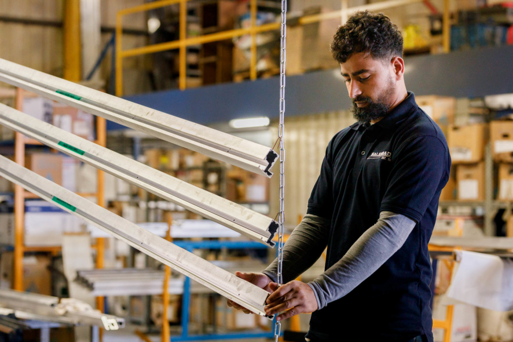 male worker in a dark blue polo shirt and grey undershirt handling long pieces of aluminium in a factory or warehouse setting